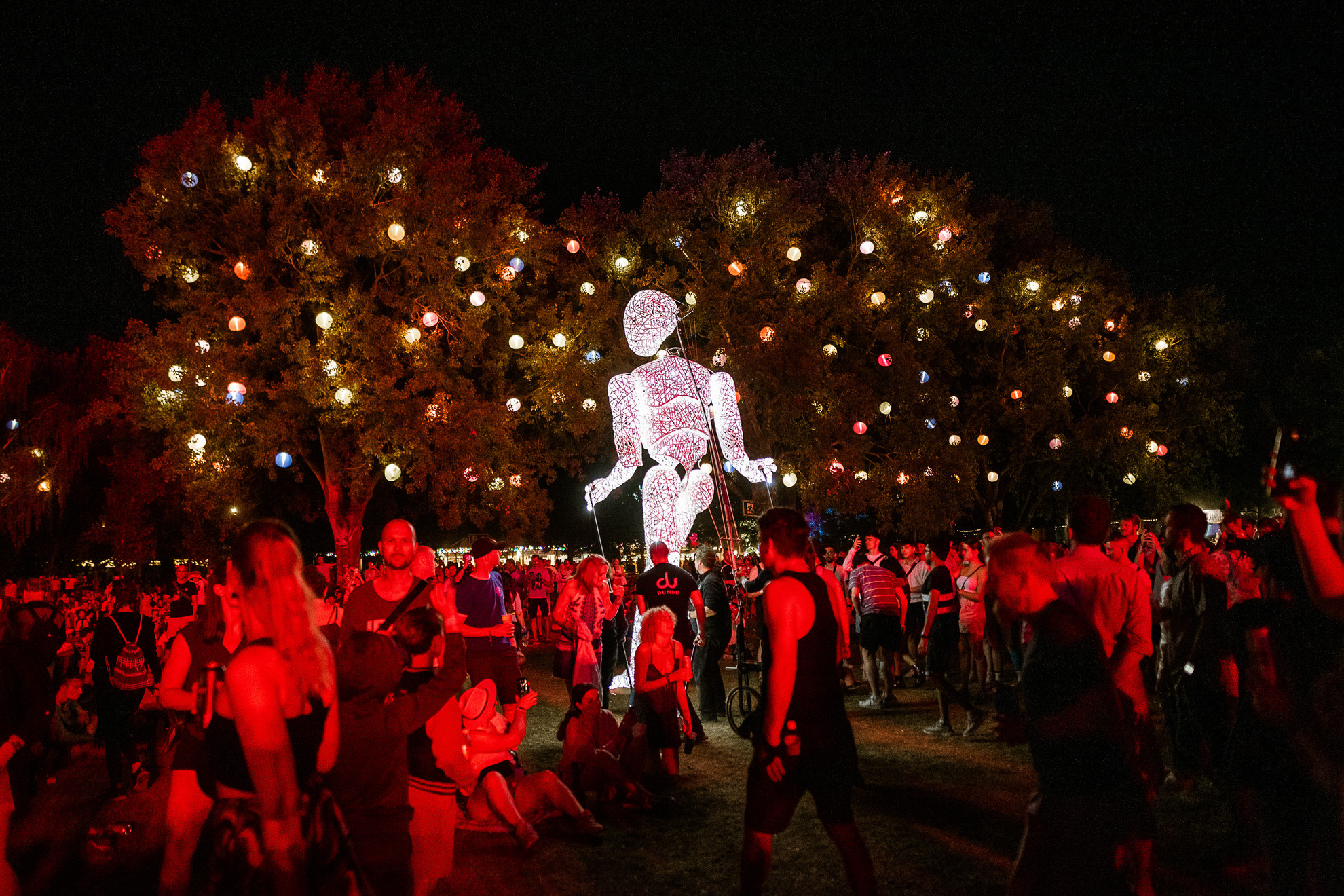 Fotó: Sziget Fesztivál / szigetfestival.com Éjszakai életkép a Sziget Fesztiválról világító óriásbábbal, színes fénygömbökkel díszített fákkal és ünneplő tömeggel, látványos utcaszínházi pillanat Budapesten.