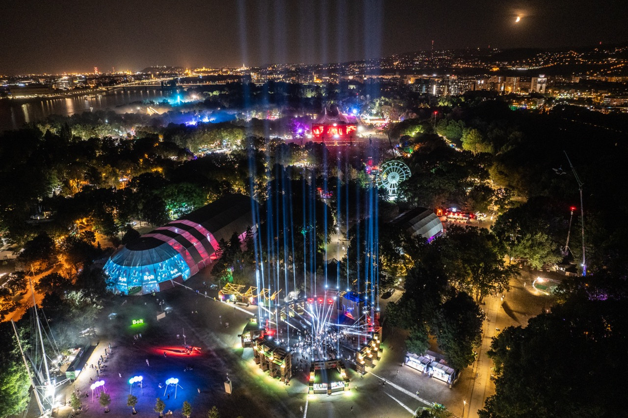 Fotó: Sziget Fesztivál / szigetfestival.com Éjszakai drónfelvétel a Sziget Fesztiválról: színpompás fények, BOLT Night Stage központi installációja, Budapest városi panorámája és a Duna fényei egy képen.